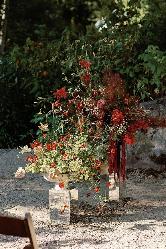 Wedding floral arrangement with red blooms, white anthurium and hanging amaranthus on a stone and acrylic pedestal in a garden setting