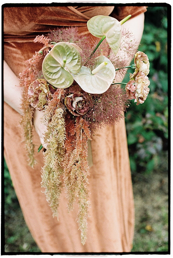 Bridal bouquet with anthurium wedding bouquet blooms, blush roses, and trailing greenery tied with ribbon against a taupe bridesmaid dress outdoors