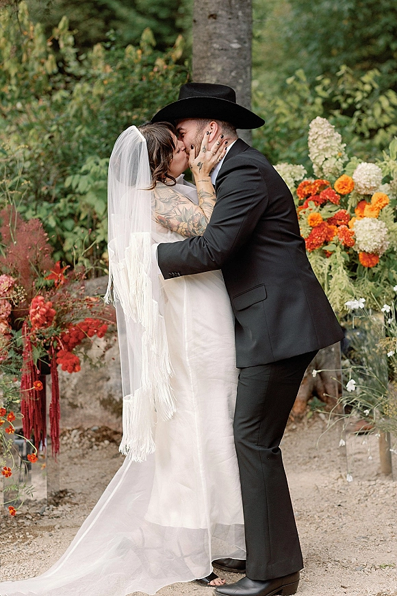 Wedding kiss portrait of bride and groom kissing as she holds his face, long veil and cowboy hat framed by garden greenery and florals