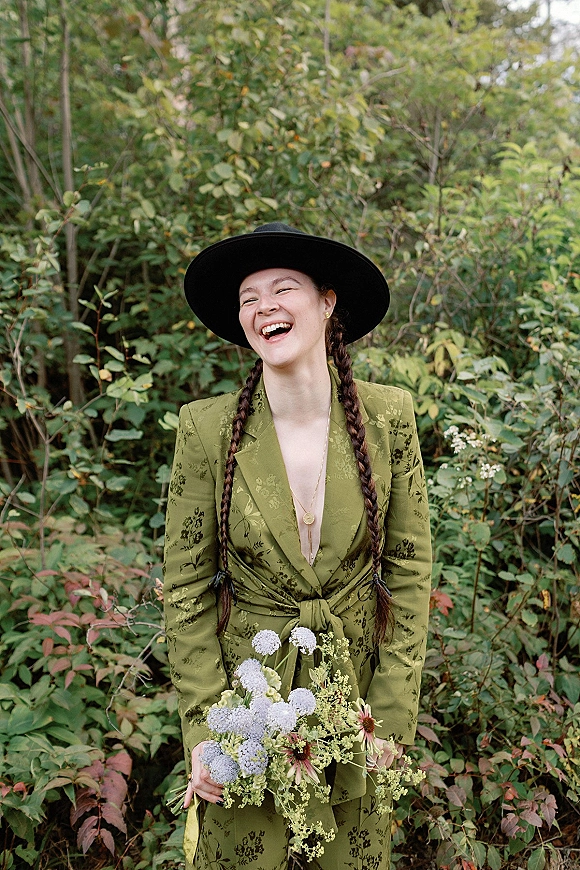 Bridal portrait of a bride wearing hat, in a green floral suit holding a wildflower bouquet, smiling among lush garden foliage