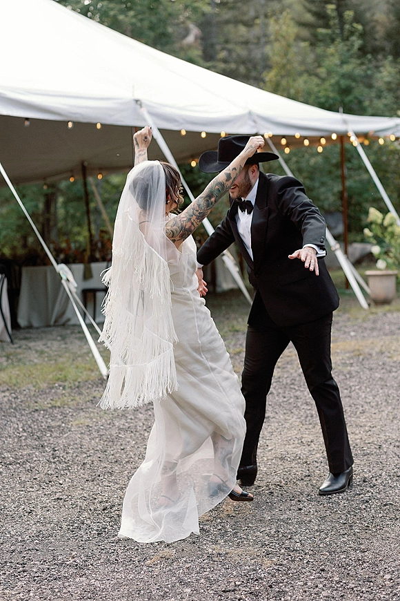 Wedding reception dancing as bride in a simple satin wedding dress and fringe veil twirls with cowboy-hat groom under string lights in an outdoor tent