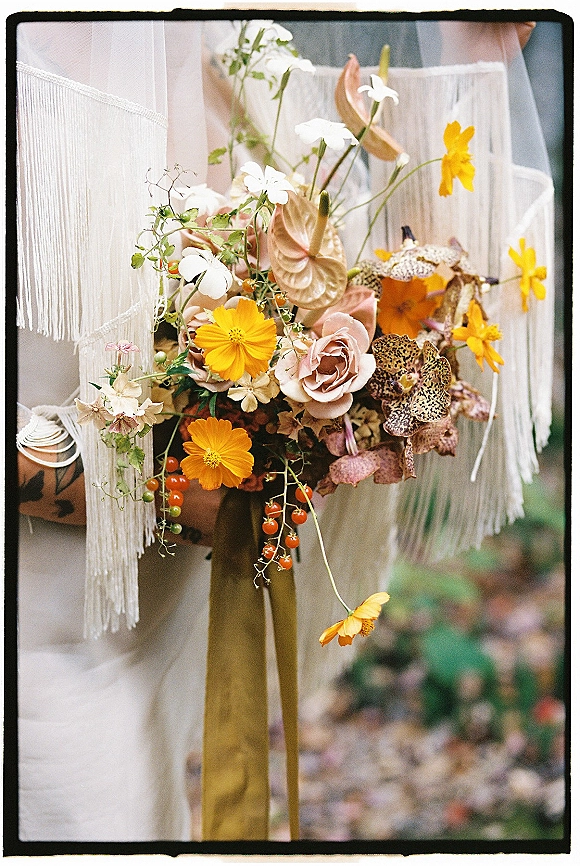 Bridal bouquet with wildflower bridal bouquet styling, blush roses, orange cosmos, and trailing ribbon wrap against blurred garden greenery