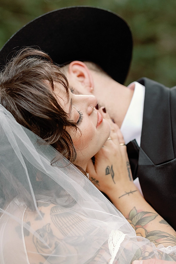 Wedding kiss as bride and groom kiss close up, her veil framing their faces, his black suit and hat against outdoor greenery