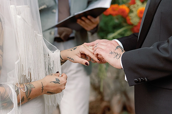 Ring exchange as bride and groom hands place wedding rings, with tattoos and veil detail during an intimate ceremony with officiant book