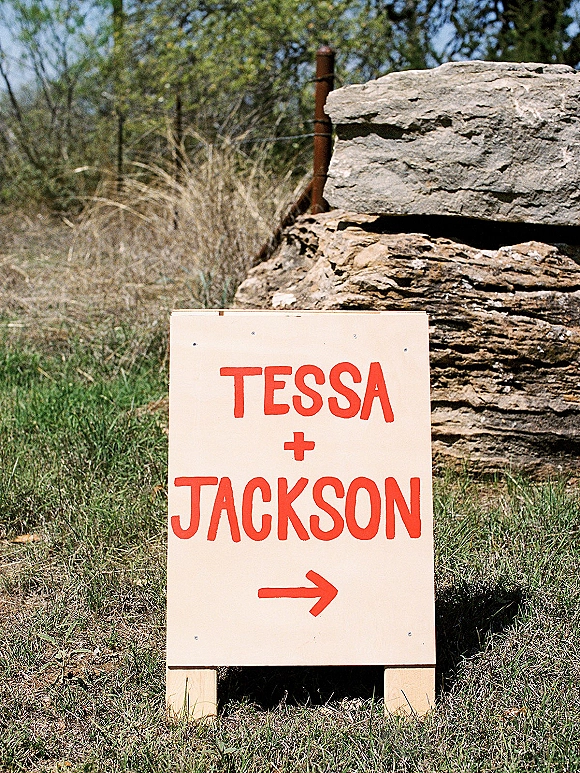 Wedding welcome sign, hand painted wedding sign with red lettering and arrow on wooden stakes in a grassy field near rocks and trees