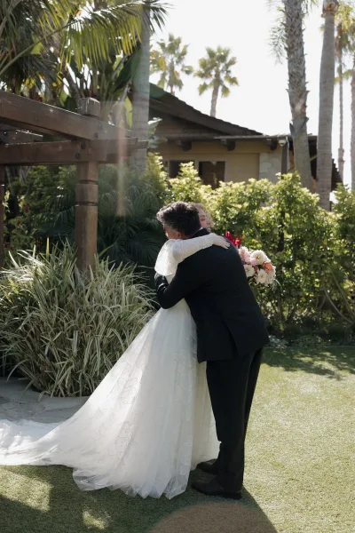 First look moment as groom hugs bride in long-sleeve gown, peach and blush bouquet in hand, on sunny lawn with palm trees and pergola