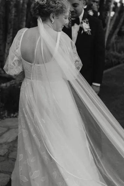 Ceremony moment as bride walks down aisle, smiling in long wedding veil and gown, groom in tuxedo on stone garden path with guests