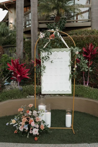 Wedding seating chart displayed on a gold metal stand with roses, greenery garland, and pillar candles in glass vases by tropical garden plants