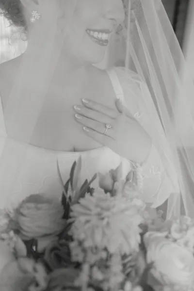 Bridal portrait in black and white of a bride smiling under her veil, hand on chest showing engagement ring in soft window light indoors