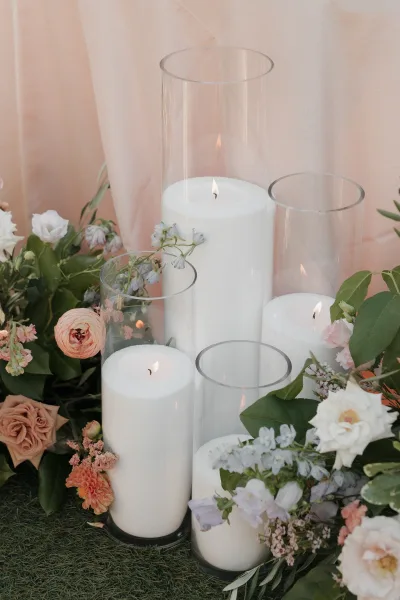 Wedding candle decor with glass hurricane candles and pillar candles in cylinder vases, nestled among florals on grass before pink drapery