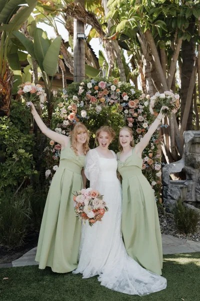 Bridesmaid portrait with bride and bridesmaids holding bouquets, arms raised beside a floral arch with tropical palms in back