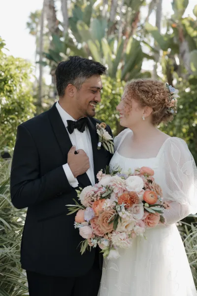 Couple portrait of bride and groom smiling shoulder to shoulder, bride holding bouquet beside groom in black tuxedo in a sunlit garden with palm trees