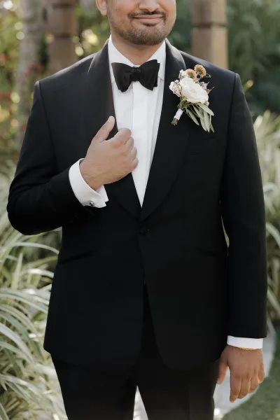 Groom portrait in a black tuxedo groom look, adjusting his jacket with boutonniere, framed by stone columns and garden greenery