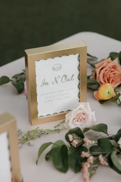 Wedding signage in a gold frame on a tabletop with rose and ranunculus florals and greenery accents against a dark green backdrop
