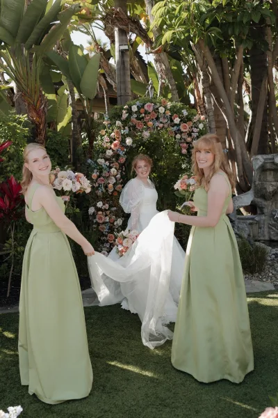 Bride with bridesmaids holding veil and bouquets in sage green dresses, standing by a floral arch amid tropical greenery and palms