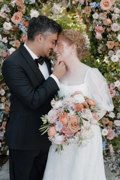 Couple portrait of bride and groom in close embrace, forehead touch, bride holding bouquet with floral wall backdrop and garden greenery