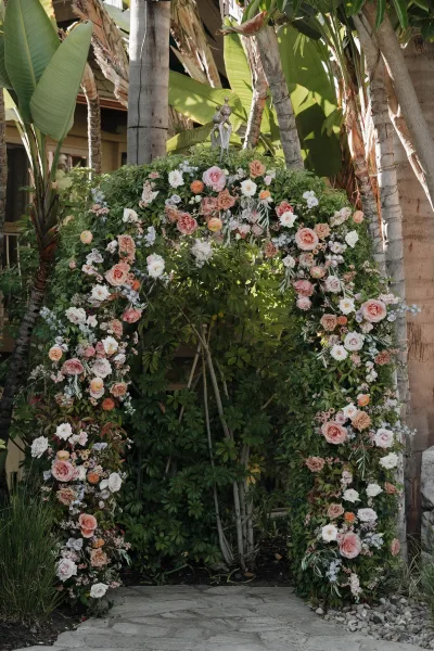 Floral wedding arch with roses and greenery, styled in pink, peach, and white blooms along a stone walkway with palm trees behind