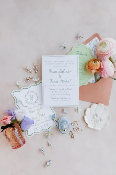 Invitation suite wedding invitation flat lay with blush envelope, wax seal, pastel flowers, rings, pearl earrings and perfume on beige fabric backdrop