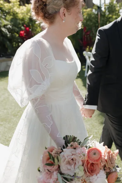 Bride portrait in side profile holding a blush and peach rose bouquet with greenery, sheer floral appliqué sleeves, in a sunlit garden with veil