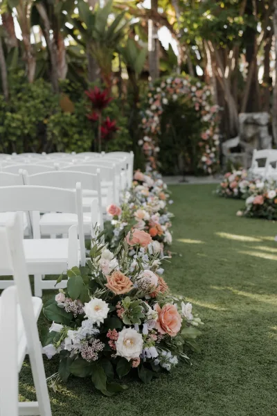 Ceremony aisle decor with blush and white rose arrangements lining white folding chairs, leading to a floral arch on a palm-lined lawn