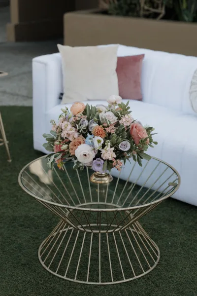 Wedding lounge decor with white sofa and throw pillows, set around a gold wire coffee table with pastel floral centerpiece on a grass lawn