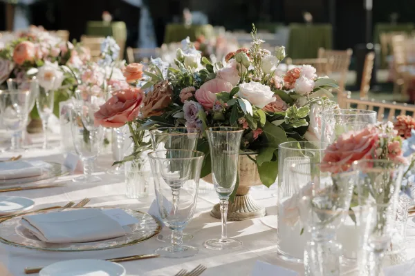 Reception tablescape with blush floral centerpiece in a stone urn, gold flatware, clear goblets, and place cards on a sunlit lawn