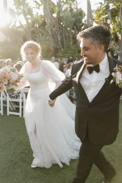 Wedding recessional as newlyweds walk hand in hand down the aisle, bride with bouquet beside groom in tuxedo, garden guests cheering
