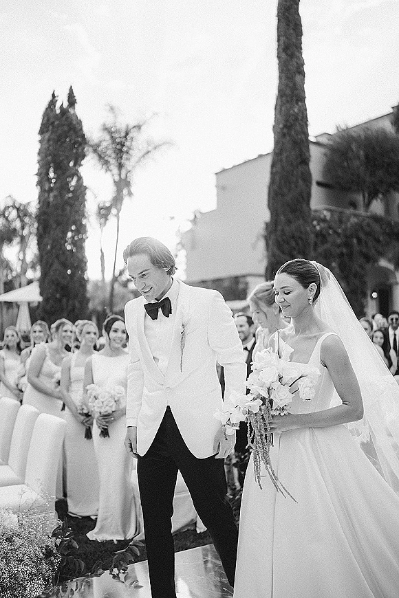 Wedding recessional as bride and groom walking aisle, bride holding bouquet and veil, guests cheering in an outdoor garden setting
