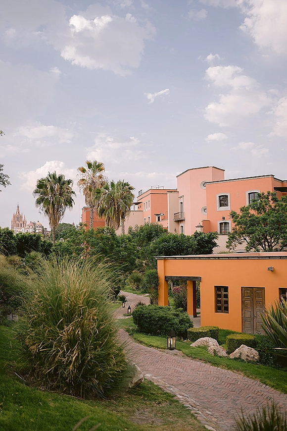 Wedding venue exterior with palm trees framing a brick walkway and pathway lanterns leading to a pastel stucco facade under clouds
