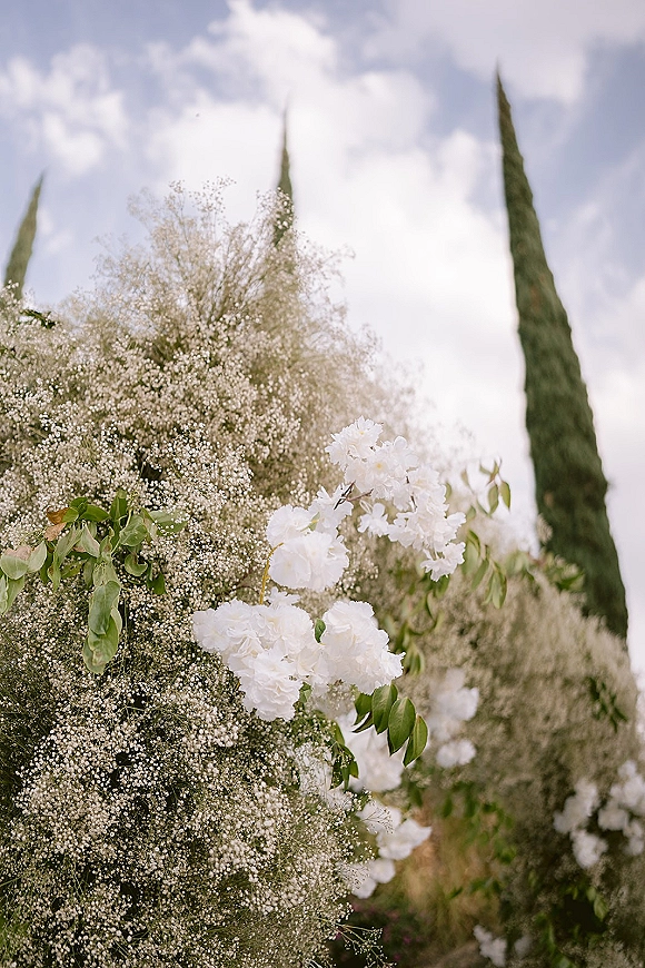 Wedding floral arrangement with baby’s breath and white flowers nestled in greenery, set outdoors beneath a blue sky with tall trees