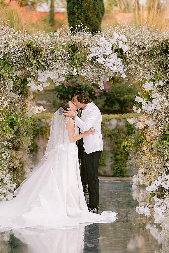 Wedding kiss beneath a white floral arch as the bride in a veil and groom in a tuxedo embrace beside a reflective garden water feature
