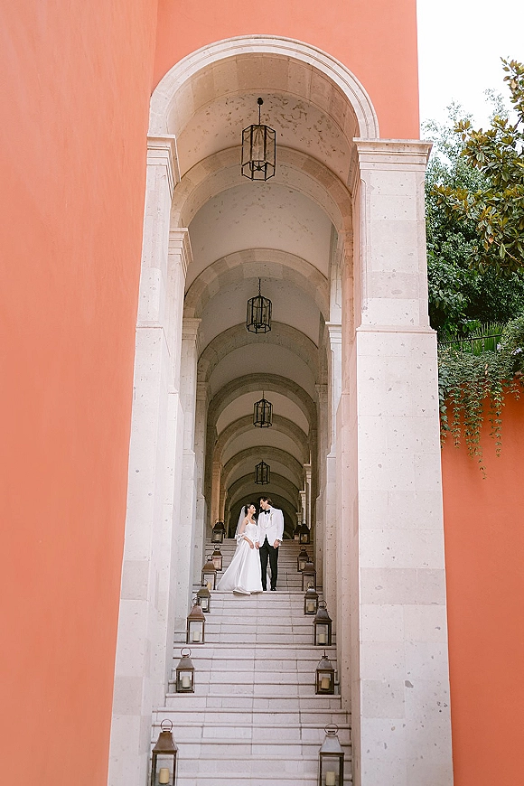 Couple portrait of bride in veil and groom in suit holding hands on a lantern-lined staircase beneath stone arches and columns
