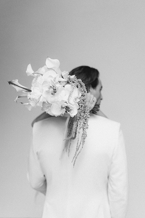 Bridal hair flowers adorn a sleek low bun with orchids and calla lilies, cascading strands beside a groom’s suit against a plain wall