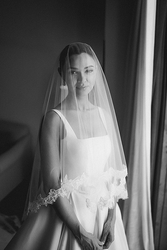 Bridal portrait in black and white of a bride with veil over her face, wearing a satin gown with lace trim in window light indoors