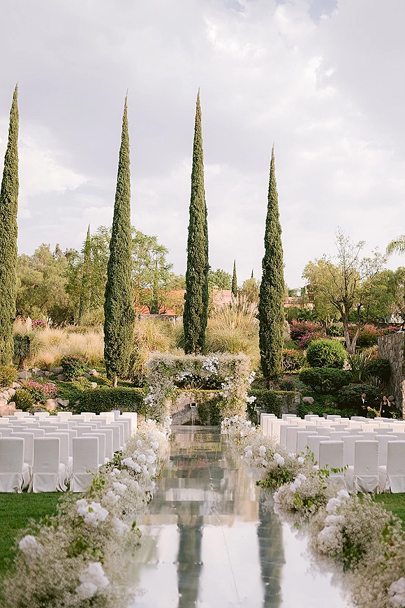 Ceremony setup with a white floral arch and mirrored aisle runner, aisle flowers and white chair covers in a garden with cypress trees