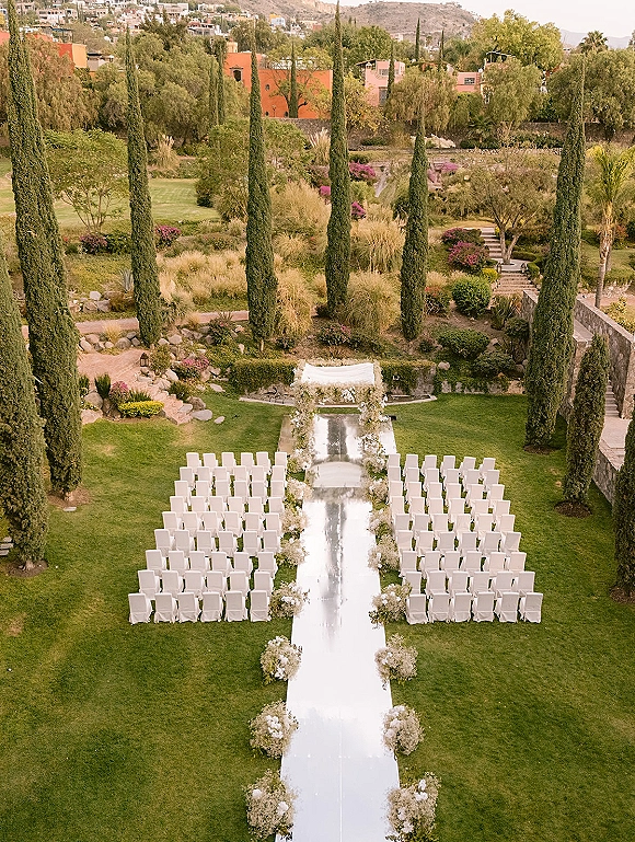 Ceremony setup with a white aisle runner leading to a floral arch, symmetrical chairs on a garden lawn with cypress trees and mountains beyond