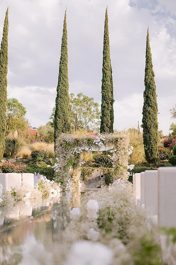 Ceremony setup with a floral arch of white flowers and greenery, white aisle runner and chairs in a garden framed by tall cypress trees