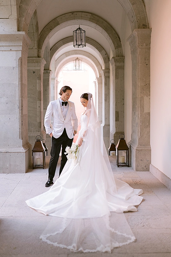 Couple portrait of bride and groom holding hands, her veil and train flowing, walking through an arched stone corridor with lanterns