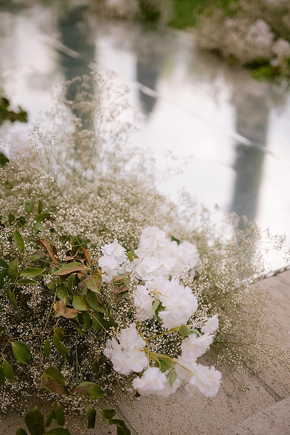 Wedding florals with baby's breath accents and white blooms arranged on stone steps beside reflective water and lush garden foliage