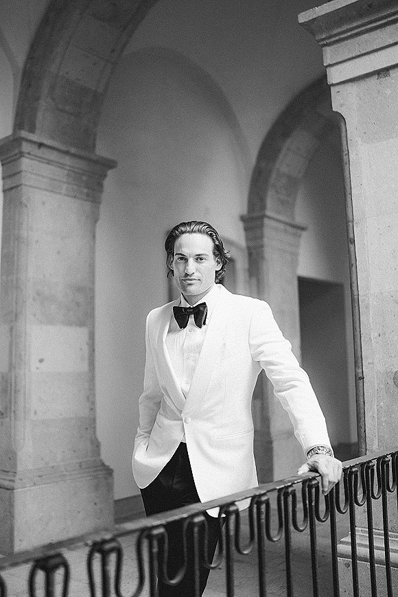 Groom portrait in a white tuxedo jacket and bow tie, leaning on a balcony railing by stone arches and columns in a courtyard hallway