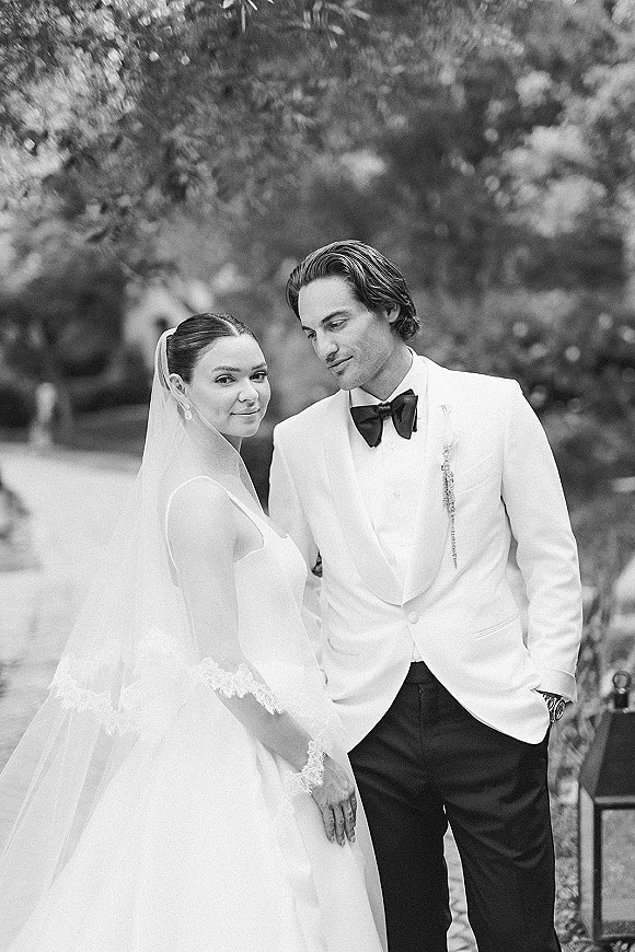 Couple portrait in a black and white wedding portrait style, bride in veil and lace sleeves with groom in tuxedo on a garden path