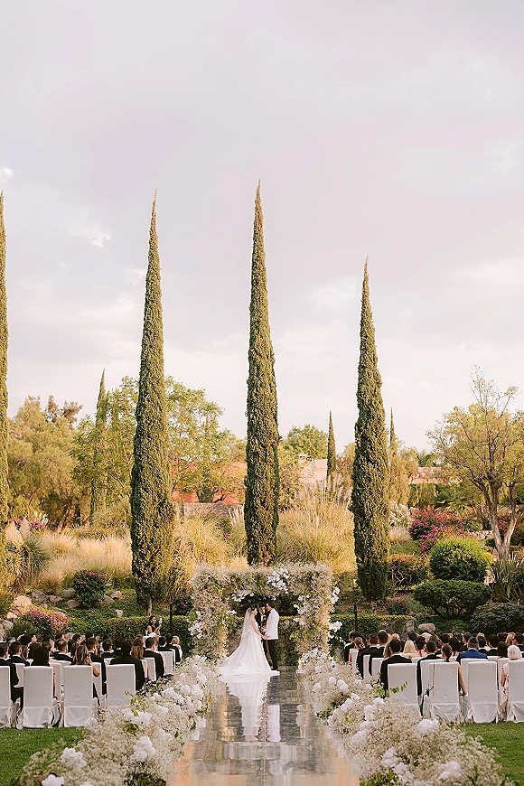 Ceremony moment as bride and groom stand at the altar during an outdoor wedding ceremony, framed by a white floral arch in a lush garden