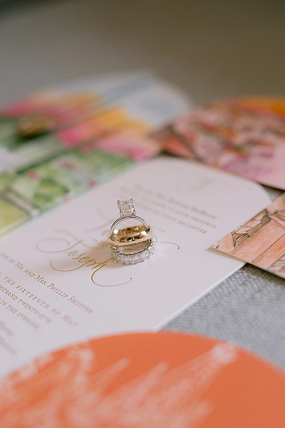 Wedding rings with an engagement ring and band resting on a gold foil calligraphy invitation card over linen, with blurred colorful paper behind