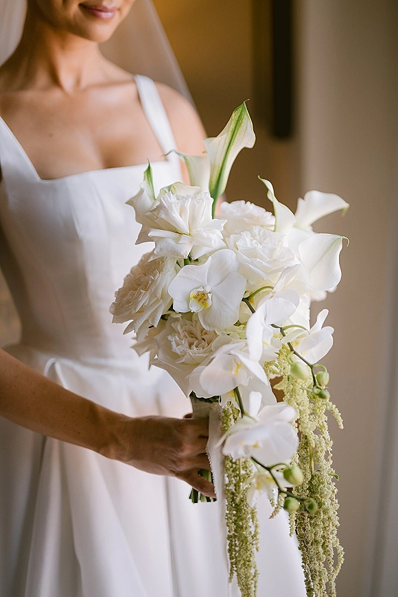 Bridal bouquet with white orchid bouquet and cascading greenery, wrapped in ribbon, held against a veil in soft window light indoors