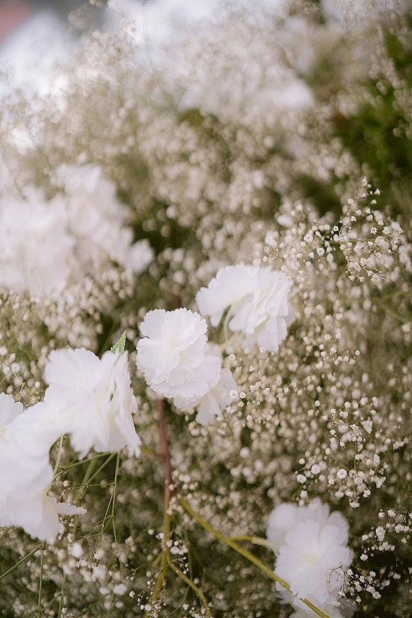 Wedding flowers of white carnations and baby’s breath with greenery stems, photographed close up against softly blurred foliage background