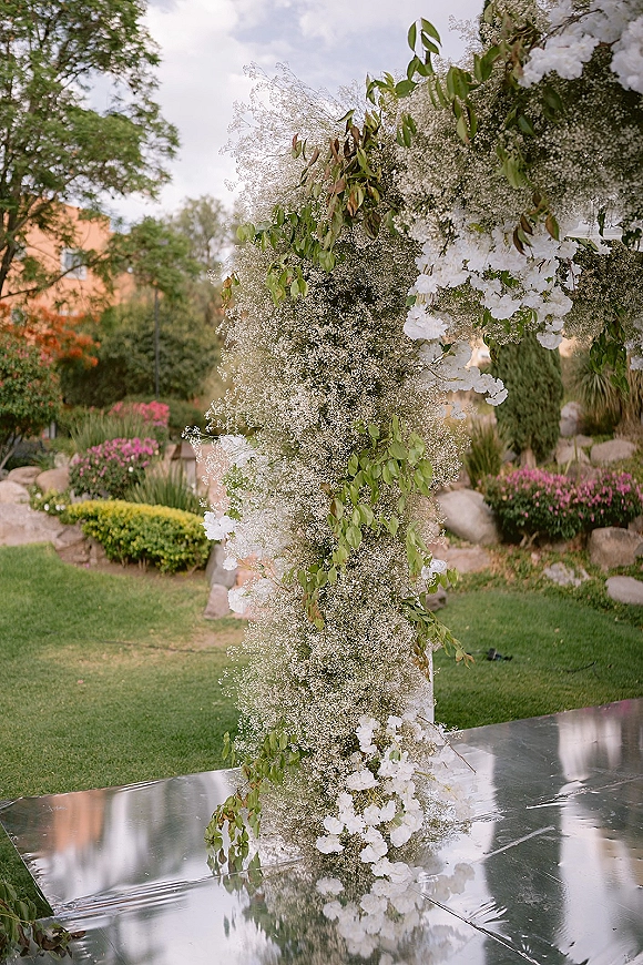 Ceremony floral arch with baby’s breath and white blooms, lush greenery and leafy branches on a reflective floor in a garden lawn setting