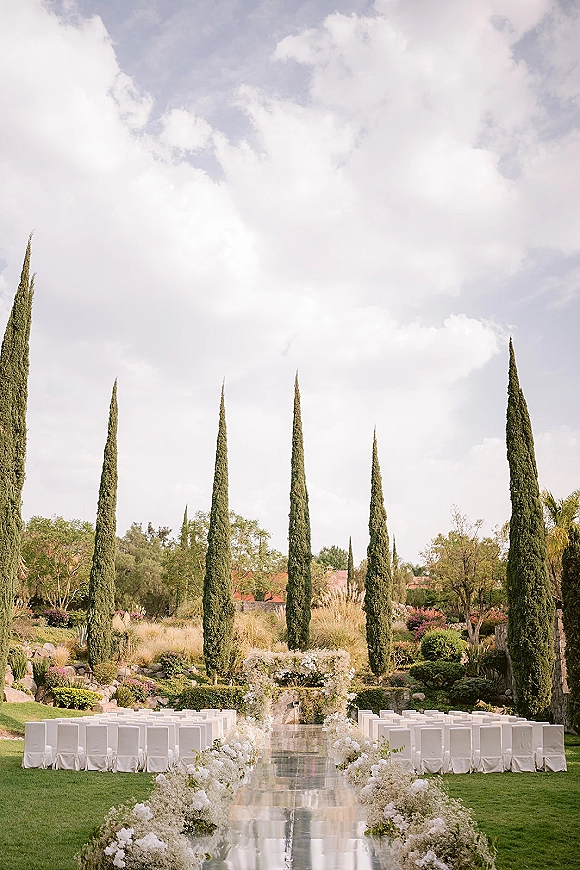Outdoor ceremony setup with clear aisle runner lined by white floral arrangements, white chairs and arch on a garden lawn under clouds