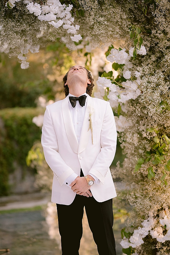 Groom portrait in a white tuxedo jacket and black bow tie, standing by a floral arch of white flowers and greenery in sunlit garden