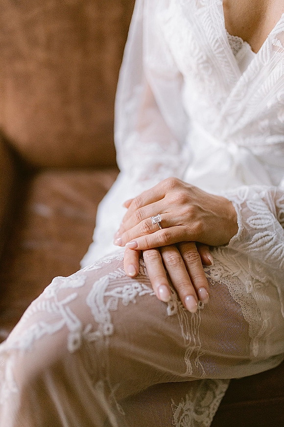 Engagement ring close-up of an emerald cut diamond on a nude-manicured hand resting on lace sleeve, with a brown leather chair behind