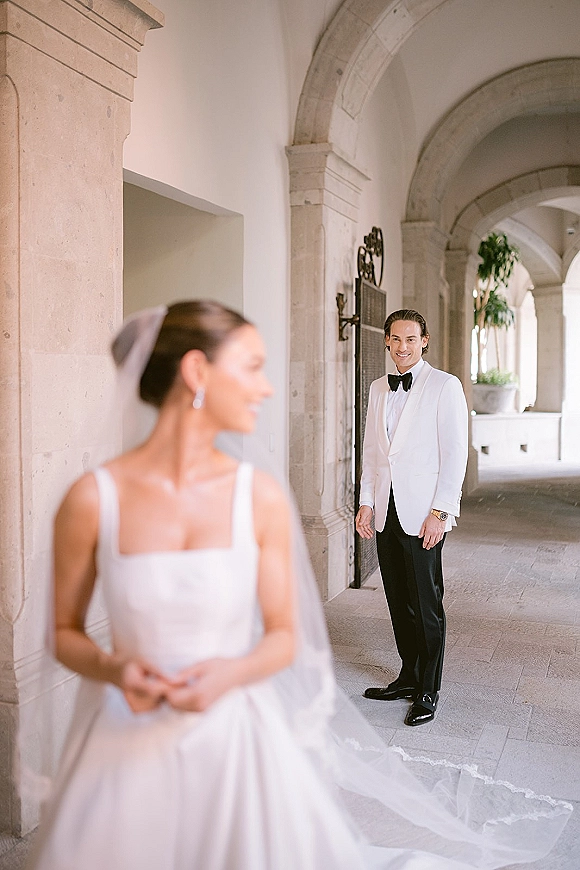 First look moment as groom in white dinner jacket sees bride in long veil and square-neck gown under a stone archway corridor